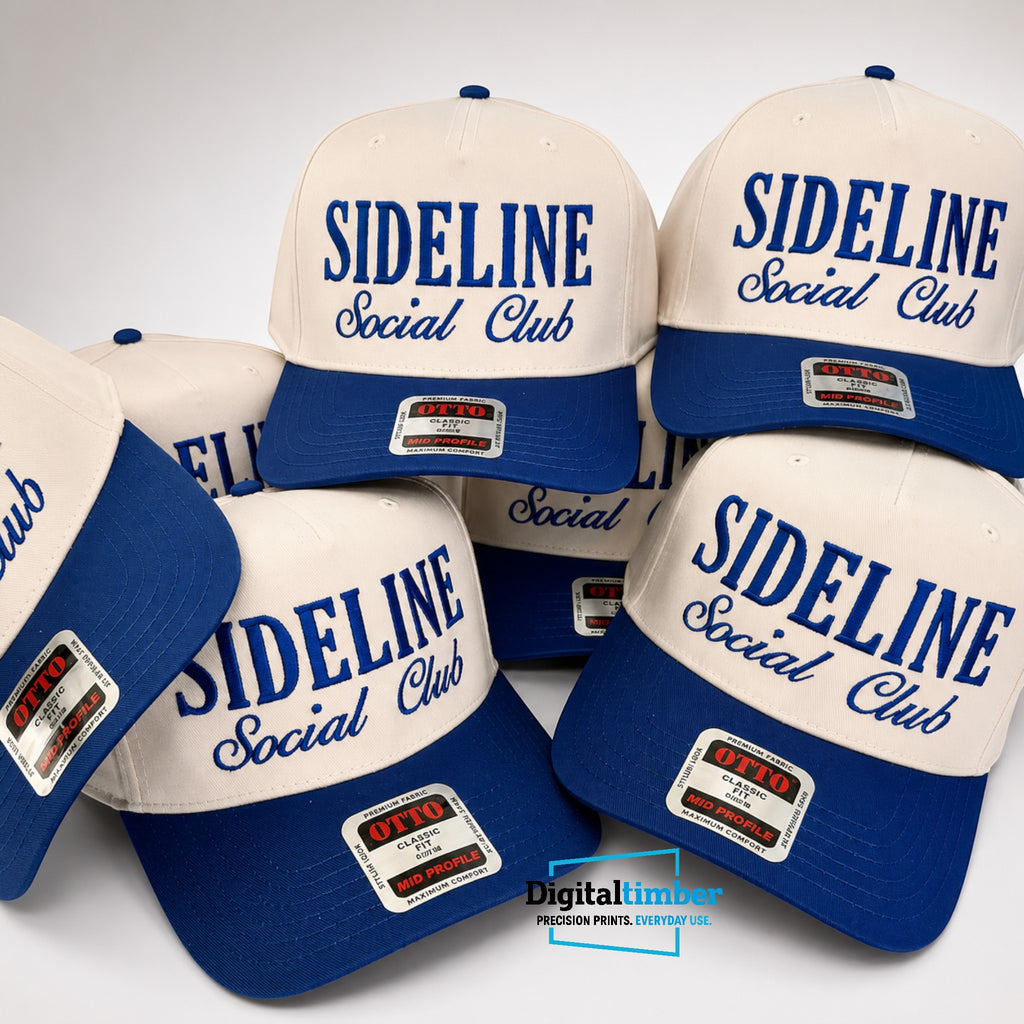 A group of baseball caps arranged together on a light gray background. The caps are off-white with royal blue visors and blue embroidered text reading 'SIDELINE Social Club.