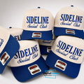 A group of baseball caps arranged together on a light gray background. The caps are off-white with royal blue visors and blue embroidered text reading 'SIDELINE Social Club.