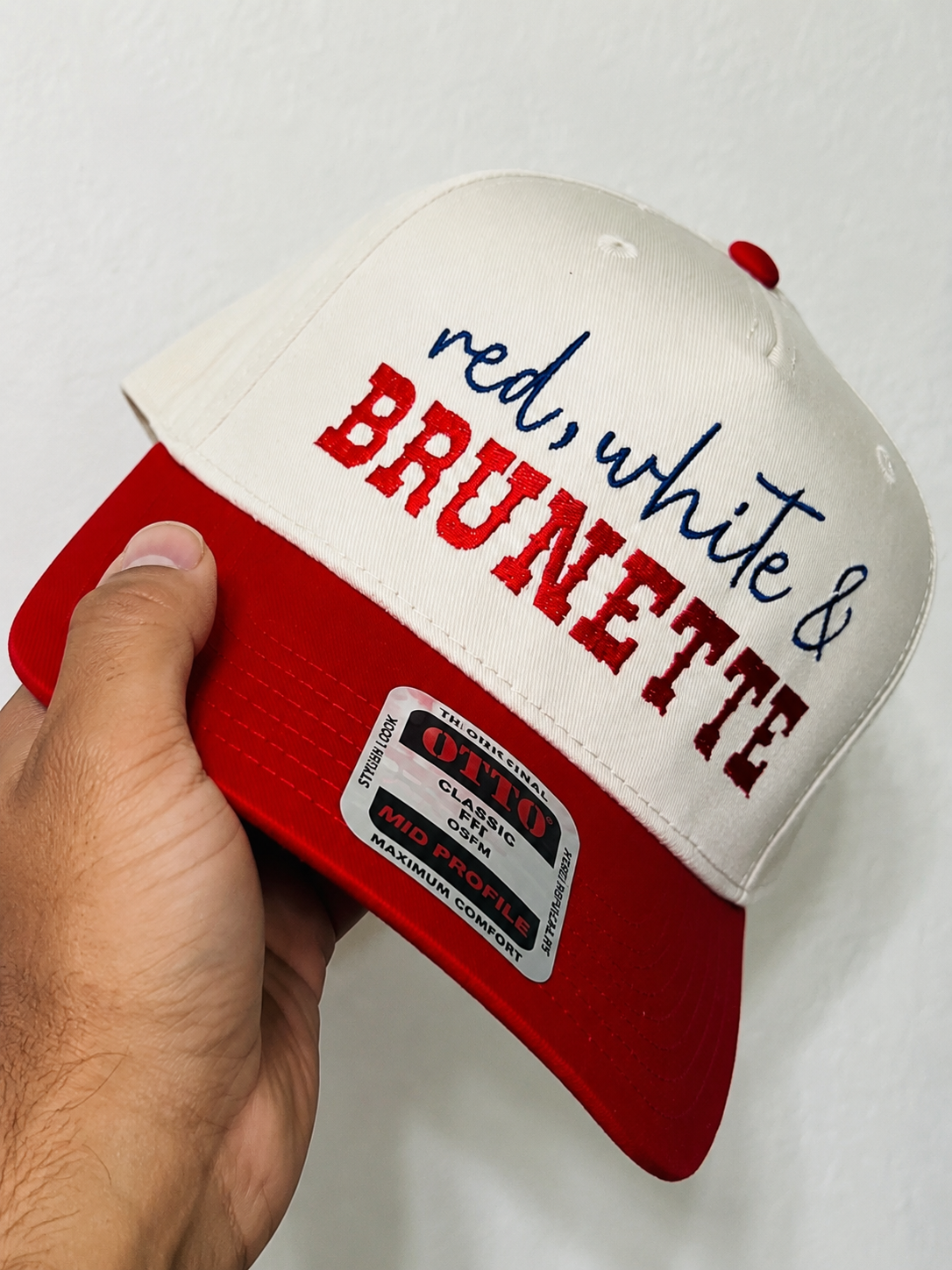 Cap with 'red, white & BRUNETTE' text held by a hand on a white background