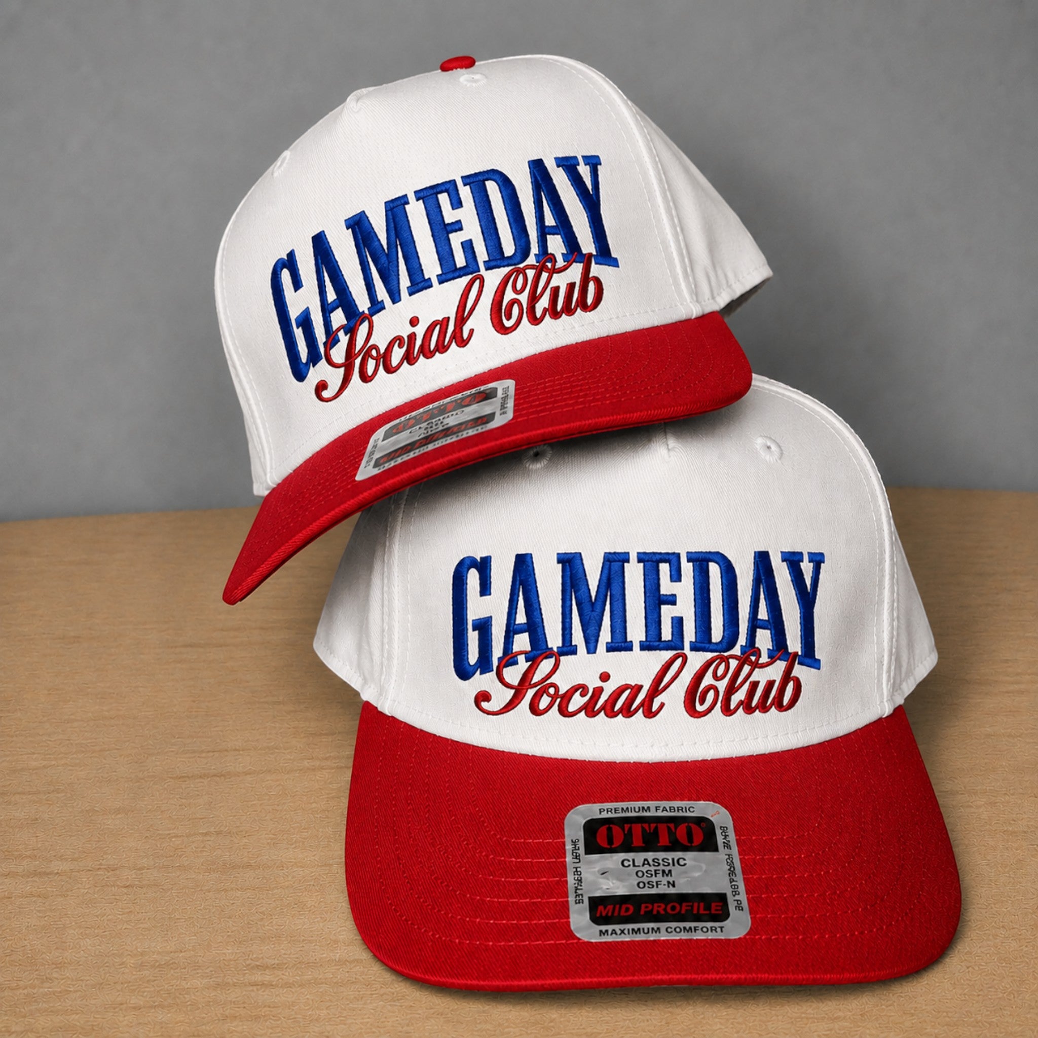 Two white and red snapback caps with 'GAMEDAY Social Club' embroidered on the front, displayed on a wooden surface against a gray background.