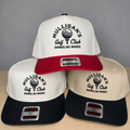 Three snapback hats arranged in a pyramid on a light wood surface against a gray background. The top hat is white with a red brim and black text reading 'MULLIGAN'S Golf Club SWINGS, NO MISSES.