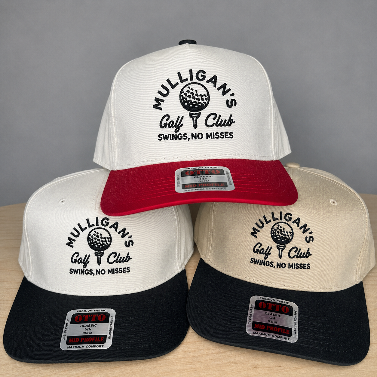 Three snapback hats arranged in a pyramid on a light wood surface against a gray background. The top hat is white with a red brim and black text reading 'MULLIGAN'S Golf Club SWINGS, NO MISSES.