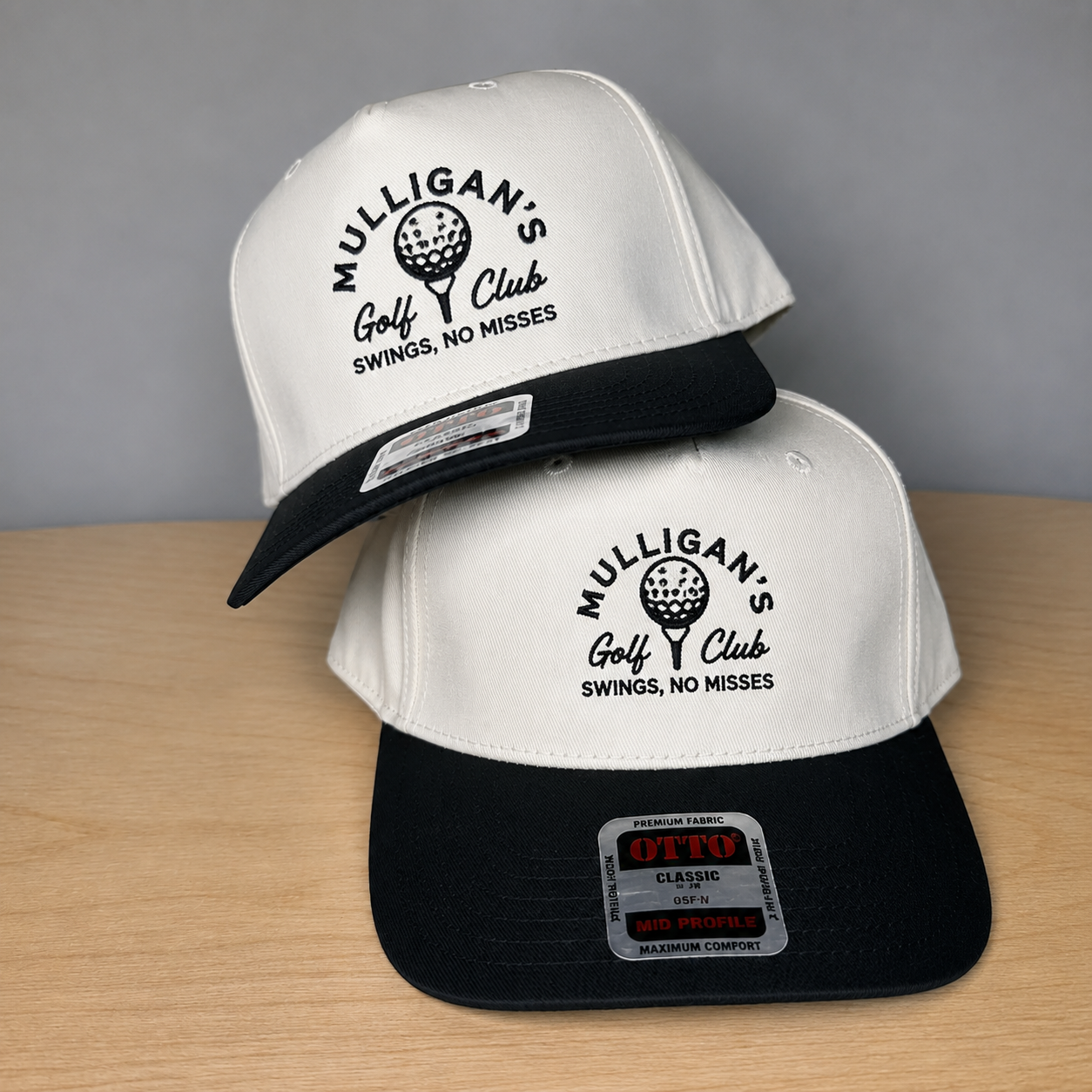 Two snapback hats stacked on a light wood surface against a gray wall. The hats are off-white on top and black on bottom, both with black curved brims and black embroidered text reading 'MULLIGAN'S Golf Club SWINGS, NO MISSES' with a golf ball graphic.
