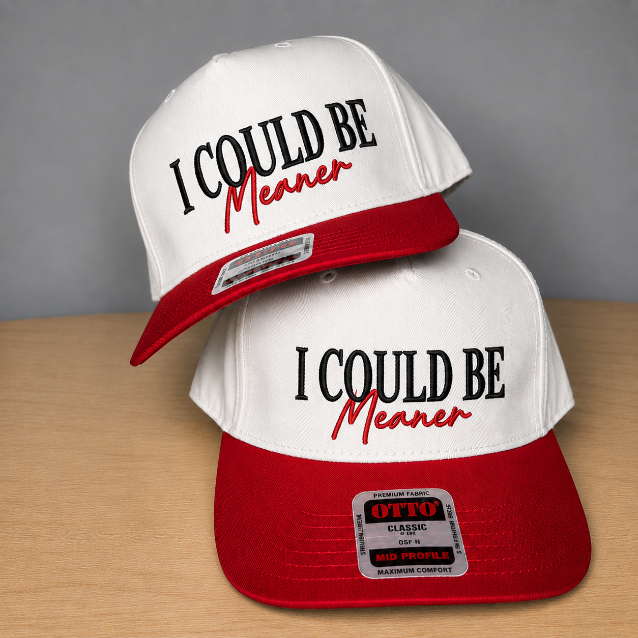Two white and red snapback caps stacked on a light wood surface against a gray background. The front of both hats reads 'I COULD BE Meaner' in black and red lettering.