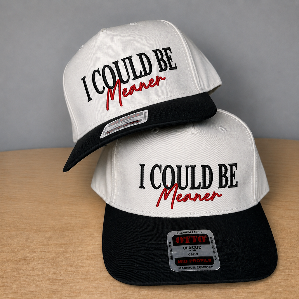 Two snapback trucker caps stacked on a light wood surface against a gray background. The caps are white on top with black brims and black/red embroidered text reading 'I COULD BE Meaner.