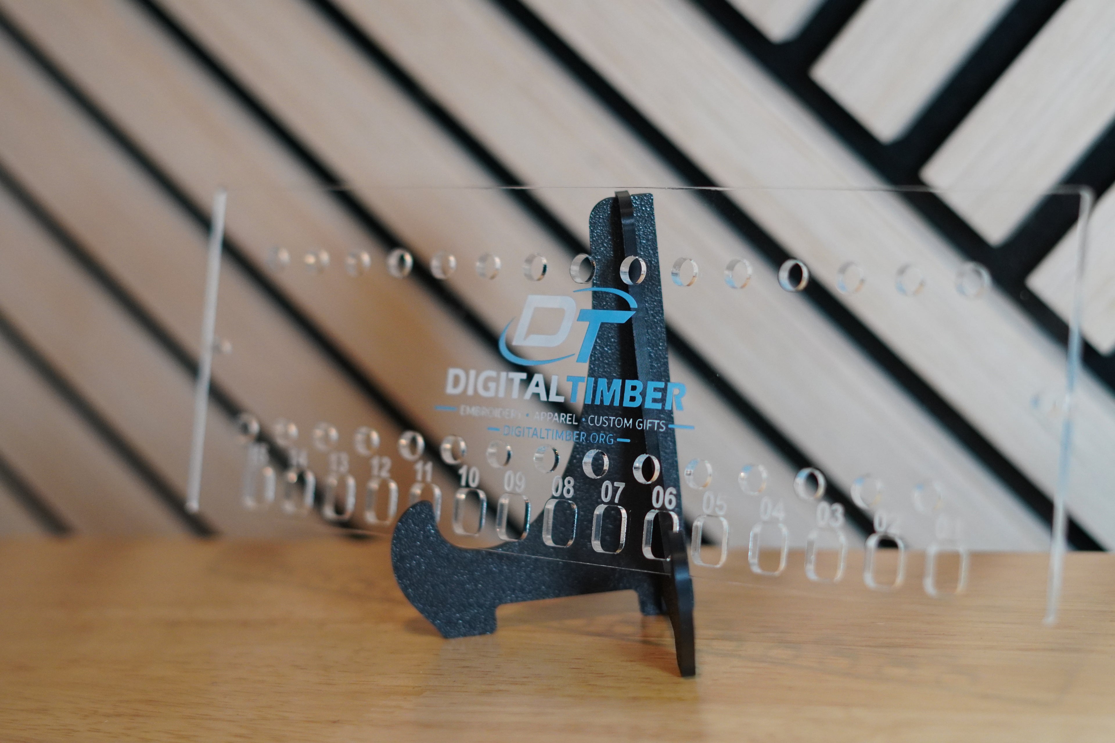 A clear acrylic digital timber gauge stand on a wooden surface, with a black digital dial and blue DIGITALTIMBER branding visible on the transparent ruler. The background shows a blurred black-and-wood geometric wall.