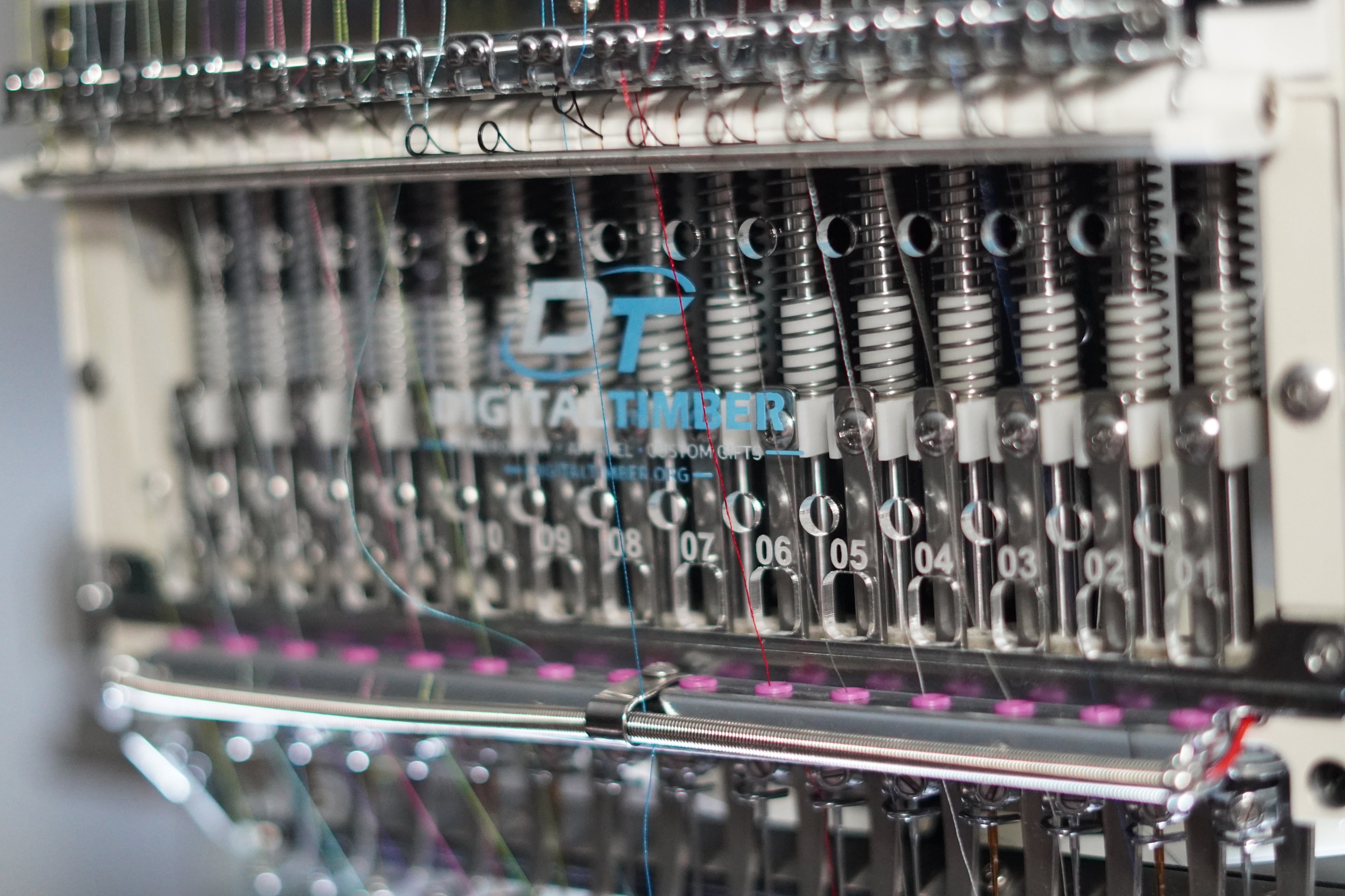 Close-up of a digital embroidery machine head with multiple needle frames, metallic components, and colorful thread lines. The brand name 'Digital Timber' is visible on the front.