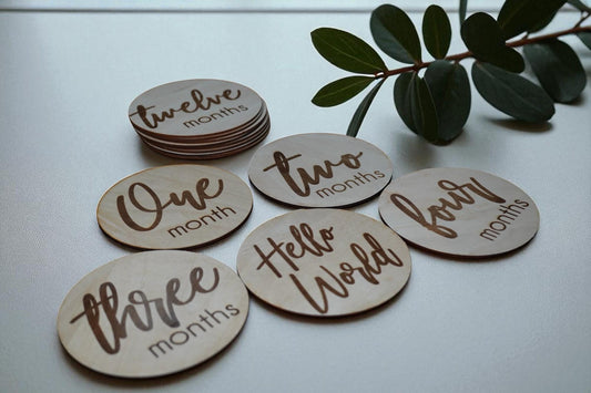 Set of engraved wooden baby milestone discs with modern script font, one through twelve months, styled with greenery for baby photography props.