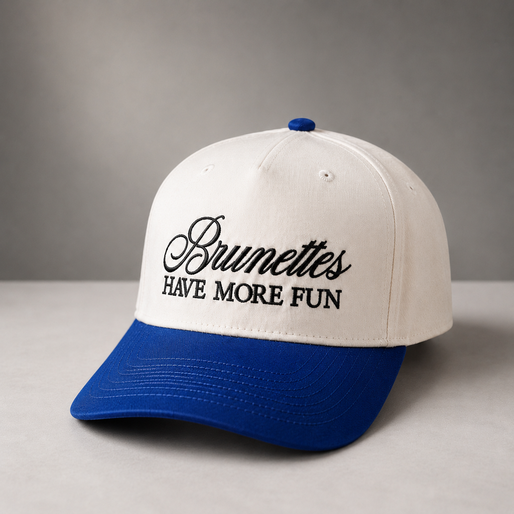 A two-tone baseball cap with an off-white crown and a royal blue curved brim. The front of the crown features black embroidered text reading 'Brunettes HAVE MORE FUN'.