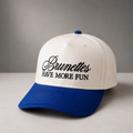 A two-tone baseball cap with an off-white crown and a royal blue curved brim. The front of the crown features black embroidered text reading 'Brunettes HAVE MORE FUN'.