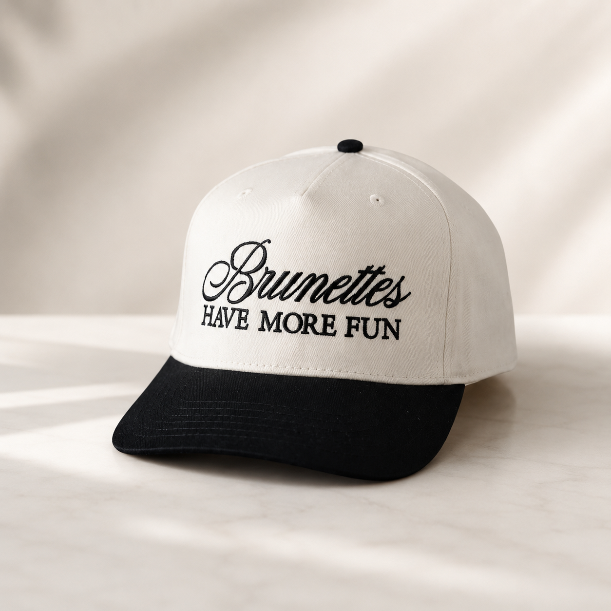 A two-tone baseball cap with an off-white crown and a black curved brim, featuring the embroidered text 'Brumettes HAVE MORE FUN' on the front. The cap is photographed on a light marble surface with soft, warm shadows.