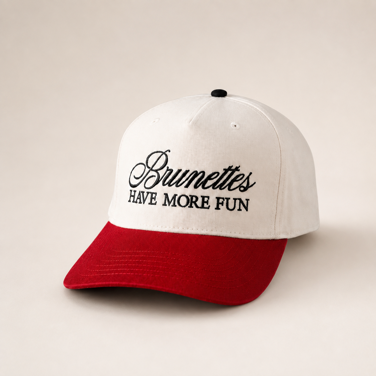 A two-tone baseball cap with an off-white crown and a red visor, featuring the embroidered text 'Brumettes HAVE MORE FUN' in black on the front. The cap is photographed on a clean, solid, warm beige background.