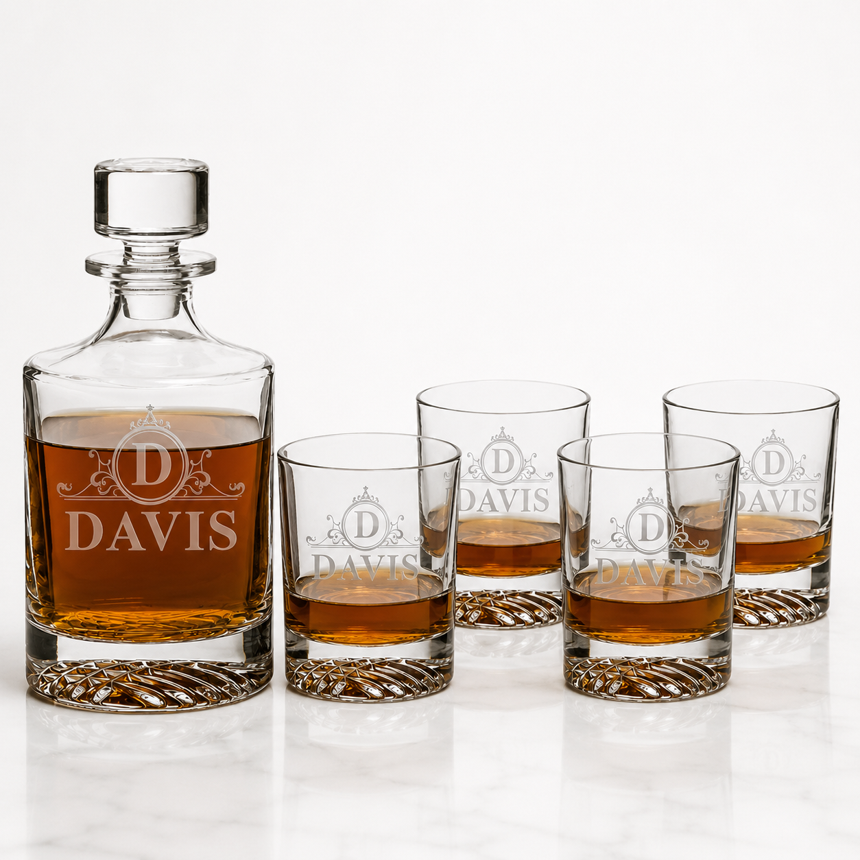 A personalized whiskey decanter and matching four rocks glasses set on a white marble surface against a white background. The decanter and glasses are made of clear glass, featuring an etched monogram 'D' with the name 'DAVIS' and a small crown motif, and filled with amber whiskey.