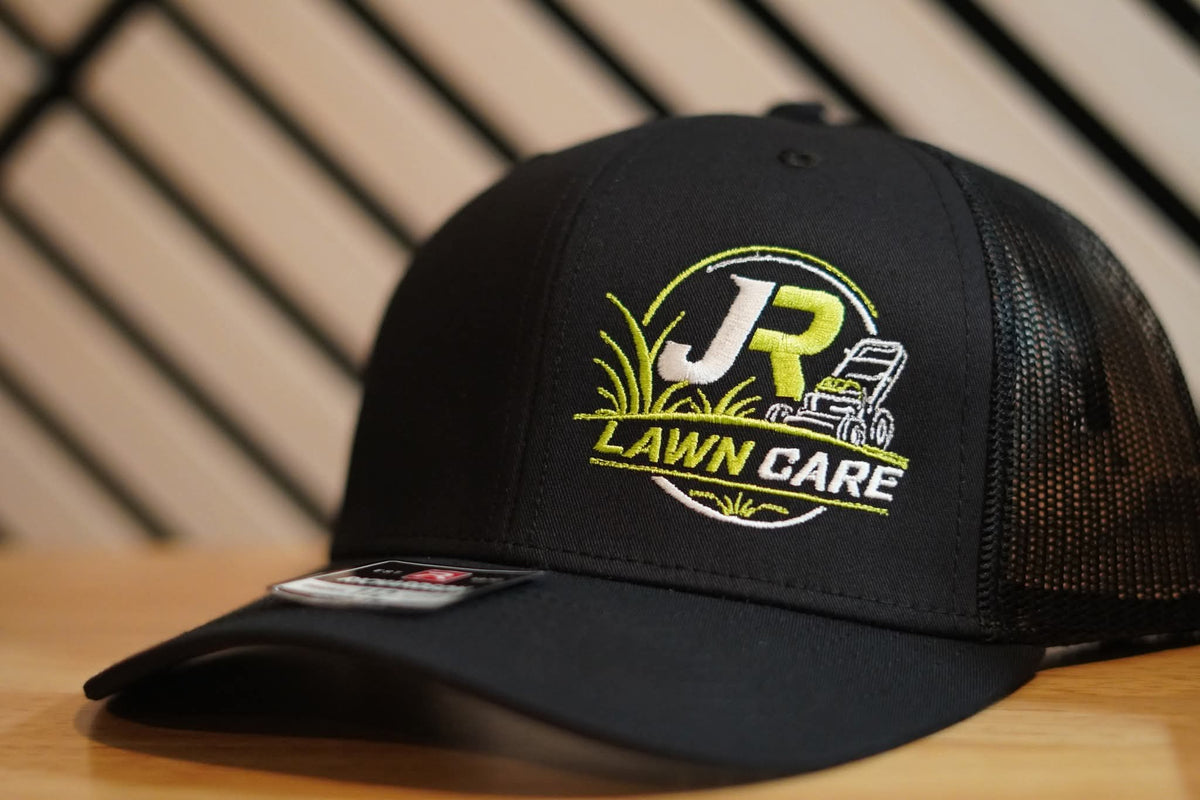 Close-up of green and white JR Lawn Care embroidery on a black trucker hat