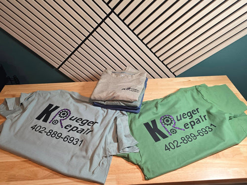 Two folded graphic T-shirts displayed on a wooden table, one light gray and one sage green, with a smaller folded shirt stacked on top. The shirts feature a large black 'Kueger Repair' logo with a purple gear graphic and the phone number '402-889-6931'.