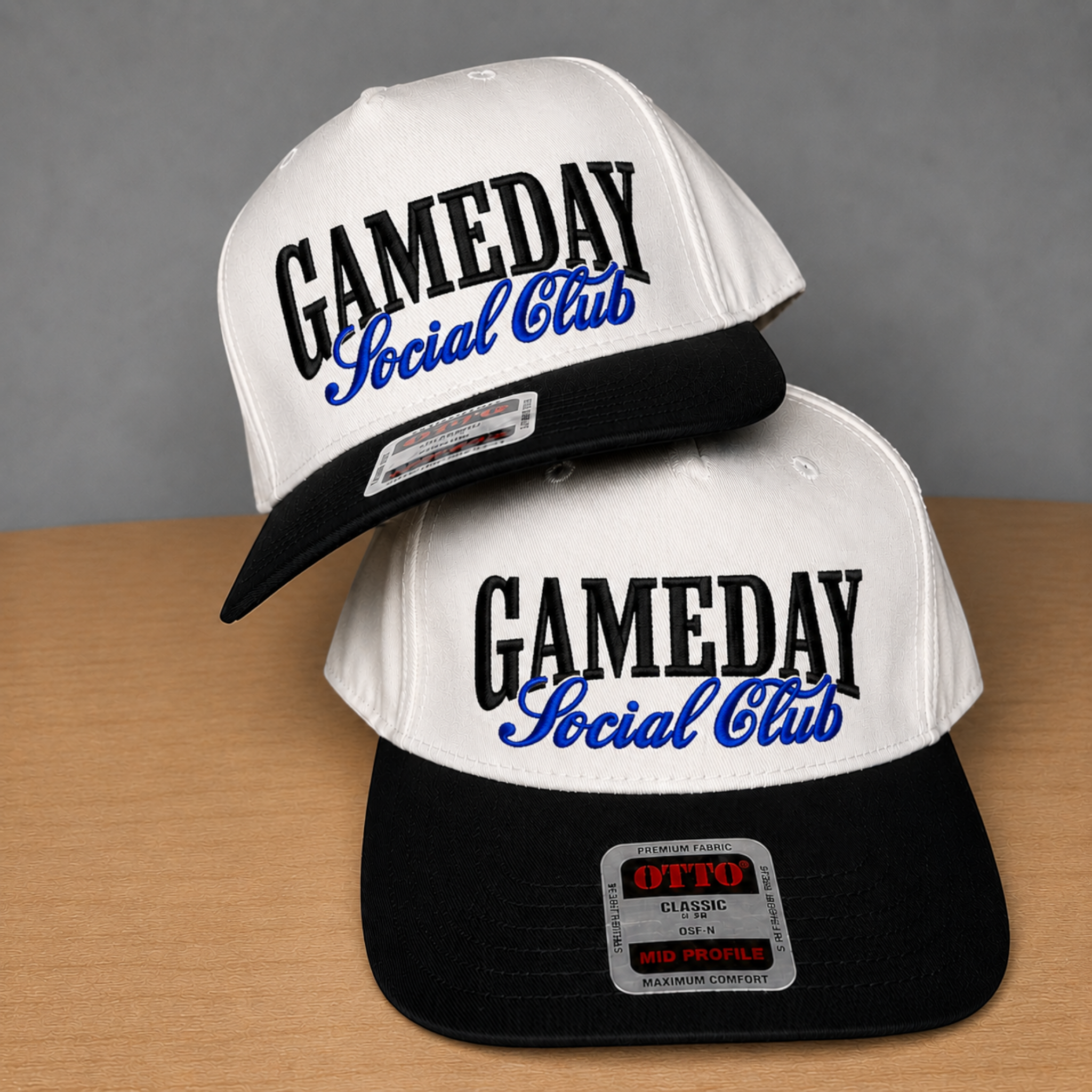 Two snapback caps stacked on a wooden surface against a gray wall. The caps are white on top with black brims and black lower panels, featuring the text 'GAMEDAY Social Club' in black and blue embroidered lettering.