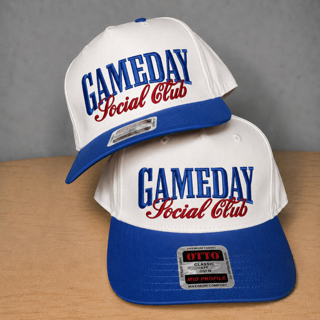 Two white and blue snapback caps with red embroidered text reading 'GAMEDAY Social Club' stacked on a wooden surface against a gray background.