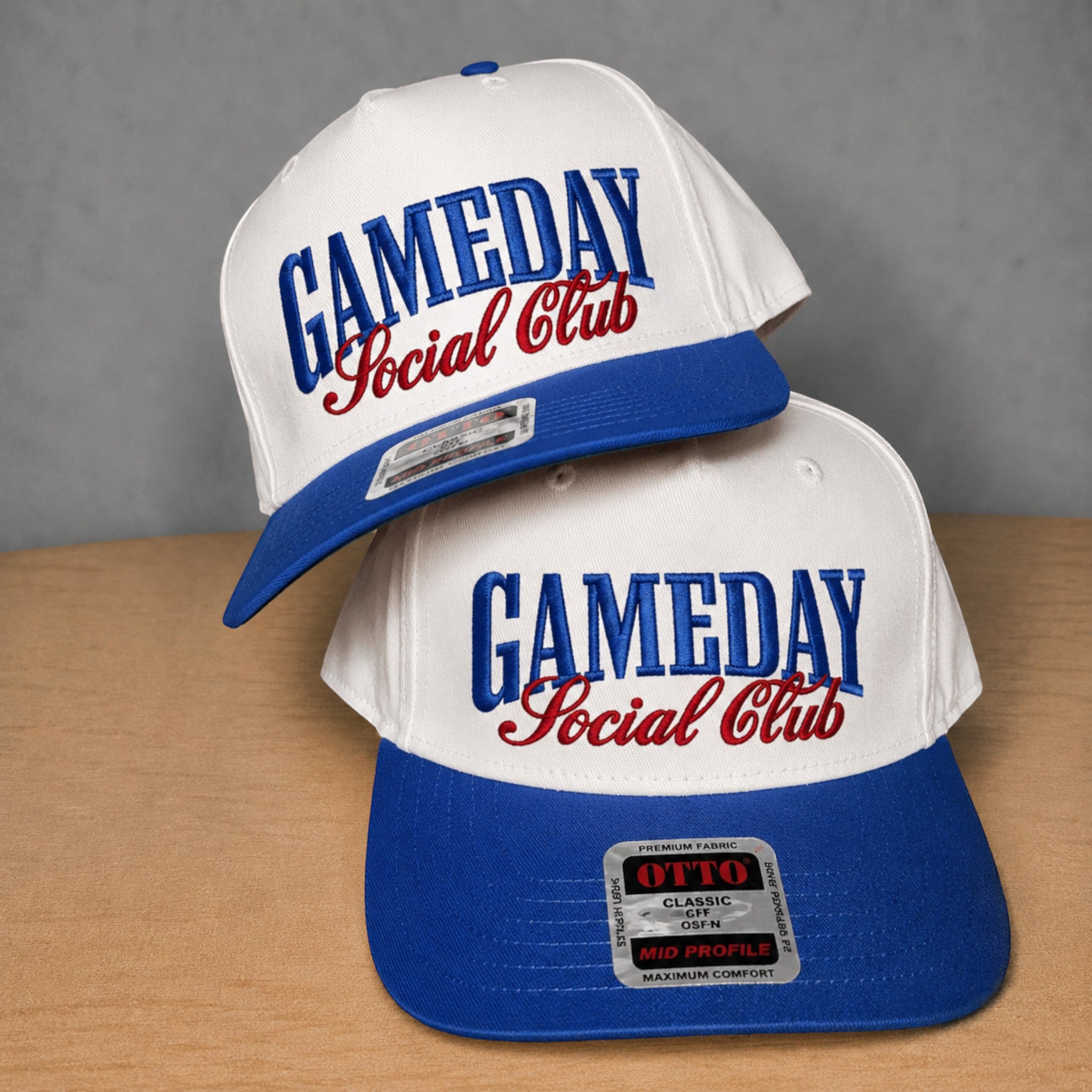 Two white and blue snapback caps with red embroidered text reading 'GAMEDAY Social Club' stacked on a wooden surface against a gray background.
