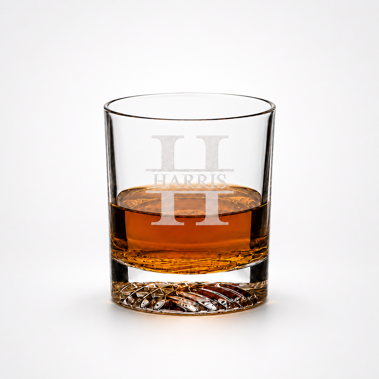A clear whiskey tumbler glass with a textured base, partially filled with amber liquor, featuring a frosted white monogram 'H H' and the word 'HARRIS' on the front, set against a clean white background.