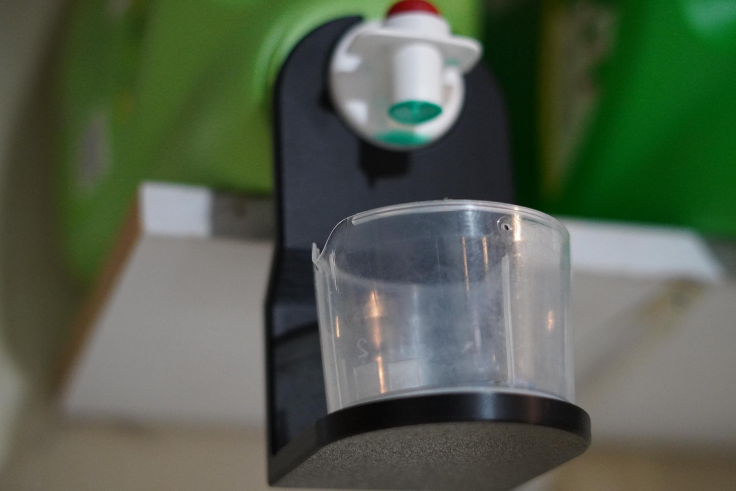 Side view of laundry detergent cup holder mounted under a jug nozzle, catching leftover drips in a clear cup. (etsy image id: 6784377067)