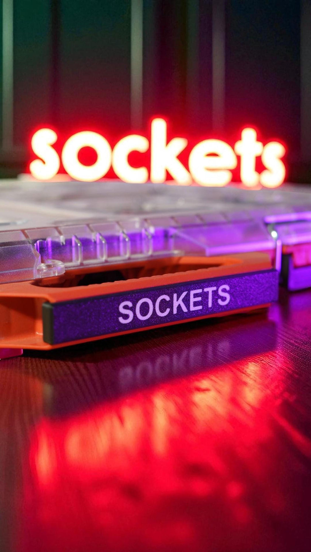 A close-up of an orange bin with a black label that says "SOCKETS" in white text, with a red neon sign in the background also spelling "sockets." __etsy:8360952149