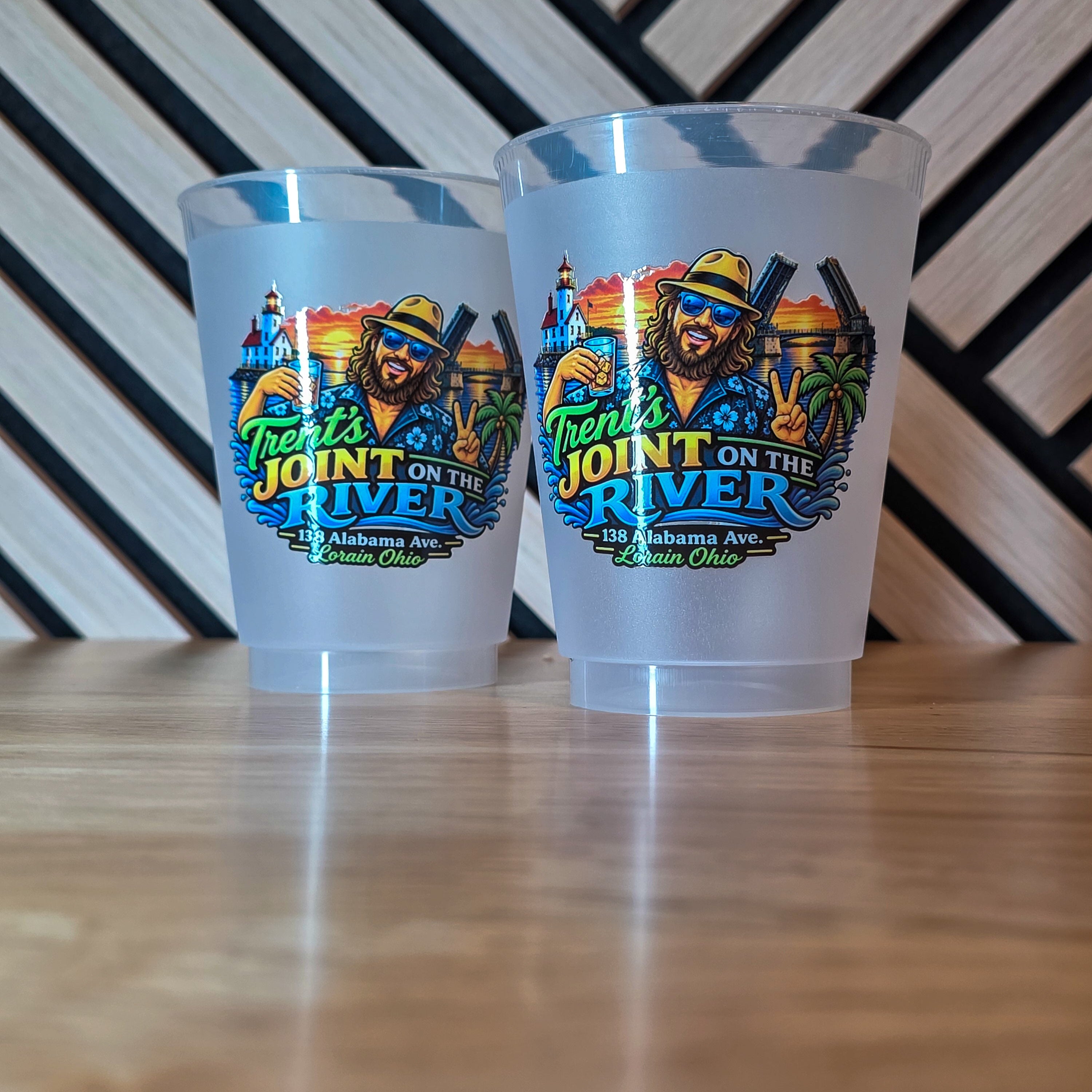 Two frosted clear plastic cups with a colorful 'Frontier's Joint on the River' graphic, displayed on a wooden tabletop against a black and white striped background.